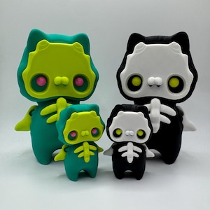 May include: Four 3D-printed cat-shaped figurines. Two are teal with yellow and pink accents, and two are black and white. The figurines have skeleton-like designs and are in varying sizes, creating a family-like display.