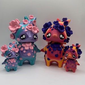May include: Four colorful, whimsical figurines with large eyes and floral accents. The figures range in size, with the largest being approximately 6 inches tall. The color palette includes pink, blue, purple, and yellow, creating a playful aesthetic.