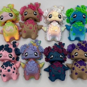 May include: A collection of twelve colorful, whimsical figurines. Each figure has a round body, small arms, and a head with large eyes and decorative floral accents. The color palette includes pink, blue, green, and brown.