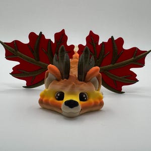 May include: A 3D-printed decorative figurine of a deer-like creature with large red maple leaf wings. The deer has a brown and orange body, small antlers, and large black eyes. The leaves have brown veins.