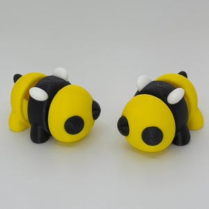 May include: Two 3D printed bumblebees, one facing left and one facing right. The bumblebees are yellow with black stripes and white wings.