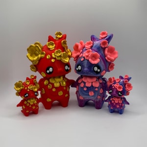 May include: Four whimsical figurines in varying sizes. Two are red with gold accents, and two are purple with pink accents. Each figure has large, expressive eyes and is adorned with flower-like details, creating a charming and artistic display.