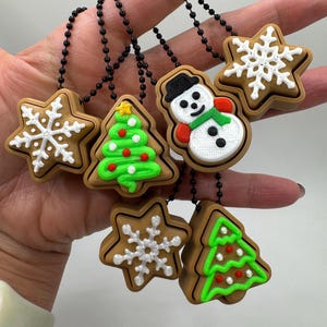 May include: A collection of Christmas-themed charms, including gingerbread-style snowflakes, Christmas trees, and a snowman. Each charm is brown with white, red, green, and black accents, and is attached to a black ball chain.