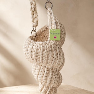 Peut inclure: Un sac à main en crochet de couleur crème en forme de coquillage en spirale. Le sac est doté d'une bandoulière épaisse tressée avec un fil doré métallique et des ferrures argentées. Une petite étiquette carrée vert clair indique Mayamay.