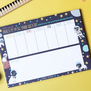 May include: A black and blue weekly planner with a space theme. It features a dog wearing a space helmet and the text "Shoot for the moon even if you miss you'll land among the stars".