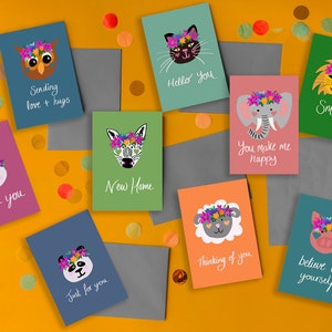 May include: A selection of greeting cards featuring different animals wearing flower crowns. The cards have various coloured backgrounds and feature messages such as "Sending love + hugs", "Hello you", "Thank you", "New Home", "Smile", "You make me happy", "Just for you", "Thinking of you", and "Believe in yourself".
