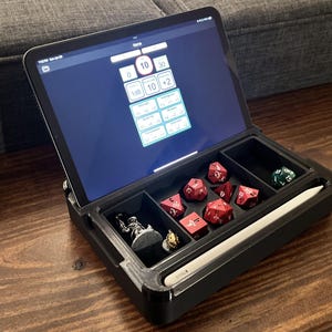Peut inclure: Une boîte noire avec un compartiment pour une tablette et un compartiment séparé pour les dés et une figurine miniature. La tablette est ouverte et affiche une application de lancer de dés numérique avec le nombre 10 affiché. La boîte comprend également un stylet.