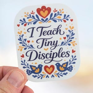 May include: A white sticker with the text "I Teach Tiny Disciples" in navy blue script. The sticker is decorated with blue leaves, red hearts, and yellow accents. Two blue birds flank a yellow heart.