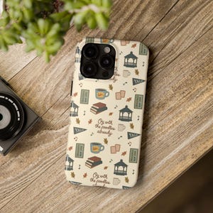 May include: A cream-colored phone case featuring a repeating pattern of dark blue gazebos, brown stacks of books, blue mugs labeled Luke's, Yale pennants, and autumn leaves. The text Oy with the poodles already is printed in a dark brown script.