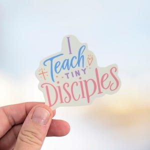 May include: A sticker with the text "I Teach Tiny Disciples" in blue, pink, and purple. The sticker has a white background and is held by a hand against a blurred background.