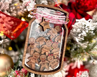 Puede incluir: Un adorno navideño de madera con forma de tarro de cristal con un inserto de vidrio transparente. El tarro está lleno de hombres de jengibre, cada uno con un nombre diferente escrito en él. Los nombres incluyen: '2021', 'Felix', 'Hugo', 'Sully', 'Elliot', 'Bailee', 'Elliodie', 'Mason', 'Papá', y 'Mamá'.