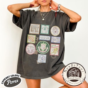 May include: Dark grey t-shirt with various stamp-like graphics. The stamps feature images and text, including "Little Piglet," "6," and "He's a Tommenbay." The shirt also has the words "Comfort Colors Pepper" and "Bendy Teez" printed on it.