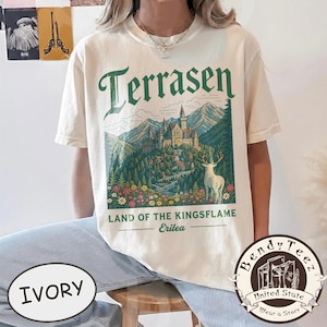 Puede incluir: Una camiseta de manga corta color marfil con un estampado gráfico de un castillo en las montañas y un ciervo blanco en primer plano. El texto dice Terrasen, Land of the Kingsflame y Erilea en letras verdes. El diseño incluye árboles y flores.