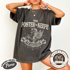 May include: Dark gray t-shirt with the text "TEAM FOSTER-KEEFE" above a winged unicorn illustration. Below the illustration is the text "WILL ALWAYS BE COOLER". The shirt has a "COMFORT COLORS PEPPER" label.