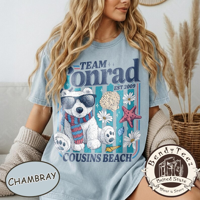 May include: Pale blue t-shirt with a graphic of a polar bear in sunglasses and a scarf, with the text "Team Conrad Cousins Beach Est 2009". The design includes seashells, starfish, and daisies.