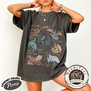 May include: Dark grey t-shirt with a colourful dragon wing graphic. The design includes various dragon wing types with text labels like "Sky Wings" and "Ice Wings". The shirt has a vintage look with the words "Comfort Colors" and "Pepper" on the bottom left.