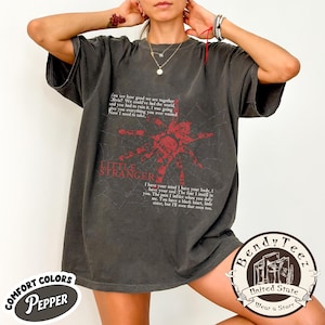 May include: Dark grey t-shirt with a distressed red spider graphic and the text "Little Stranger." The shirt also features song lyrics. The shirt has a vintage look and feel.