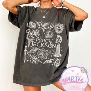 以下が含まれることがあります： 白いグラフィックデザインが施された濃いグレーのTシャツ。「Percy Jackson」の文字と、建物、ライアー、翼のある馬などのイラストが描かれています。Tシャツは半袖で、クルーネックです。