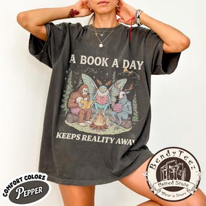 May include: A charcoal gray short-sleeve t-shirt featuring a graphic of Bigfoot, a moth creature, and a deer-headed figure sitting around a campfire reading books. The text reads A BOOK A DAY KEEPS REALITY AWAY. The shirt has a vintage-style wash.