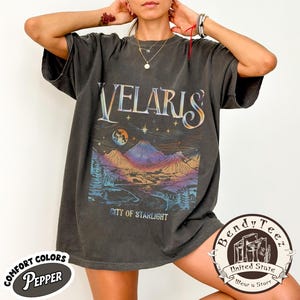Può includere: Una maglietta grigio scuro con la scritta "VELARIS" in un font stilizzato. Sotto, un paesaggio colorato con montagne, una luna e la scritta "CITY OF STARLIGHT". La maglietta ha un aspetto vintage e le parole "COMFORT COLORS PEPPER" sono stampate in basso a sinistra.