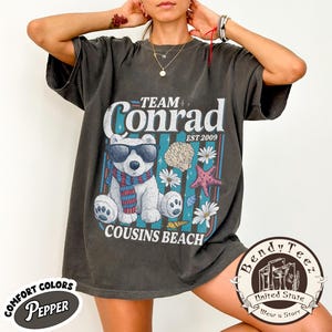 May include: Dark grey t-shirt with a graphic featuring a bear wearing sunglasses and a scarf, with the text "Team Conrad Cousins Beach Est 2009". The shirt has a vintage look and feel.