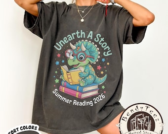 May include: Dark grey t-shirt with a graphic of a cartoon dinosaur reading a book, with the text "Unearth A Story Summer Reading 2026". The shirt also has the words "Comfort Colors Pepper" printed on it.