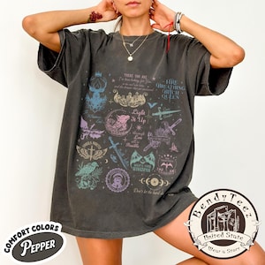 Puede incluir: Una camiseta gris carbón de corte oversize con un estampado gráfico multicolor que incluye varias ilustraciones de fantasía, como espadas, alas y textos como "Light It Up", "Umbra Mortis" y "Fire Breathing Bitch Queen" en un diseño de collage.
