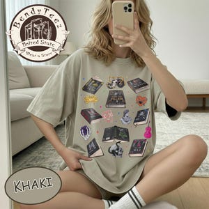 May include: Khaki-colored t-shirt featuring a collection of illustrated books with various designs and titles. The shirt includes the text "Bendy Teez" and "United State Wear a Story". The design incorporates a skull, a violin, and other graphics.