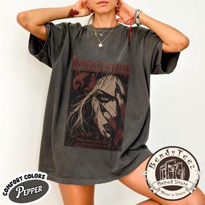 Może przedstawiać: Ciemnoszary t-shirt z krótkim rękawem z nadrukiem graficznym przedstawiającym postać z długimi włosami i ostrymi rysami twarzy. Tekst głosi Manon Blackbeak oraz From now until the darkness claims us. Koszulka ma wyblakły, stylizowany na vintage wygląd.