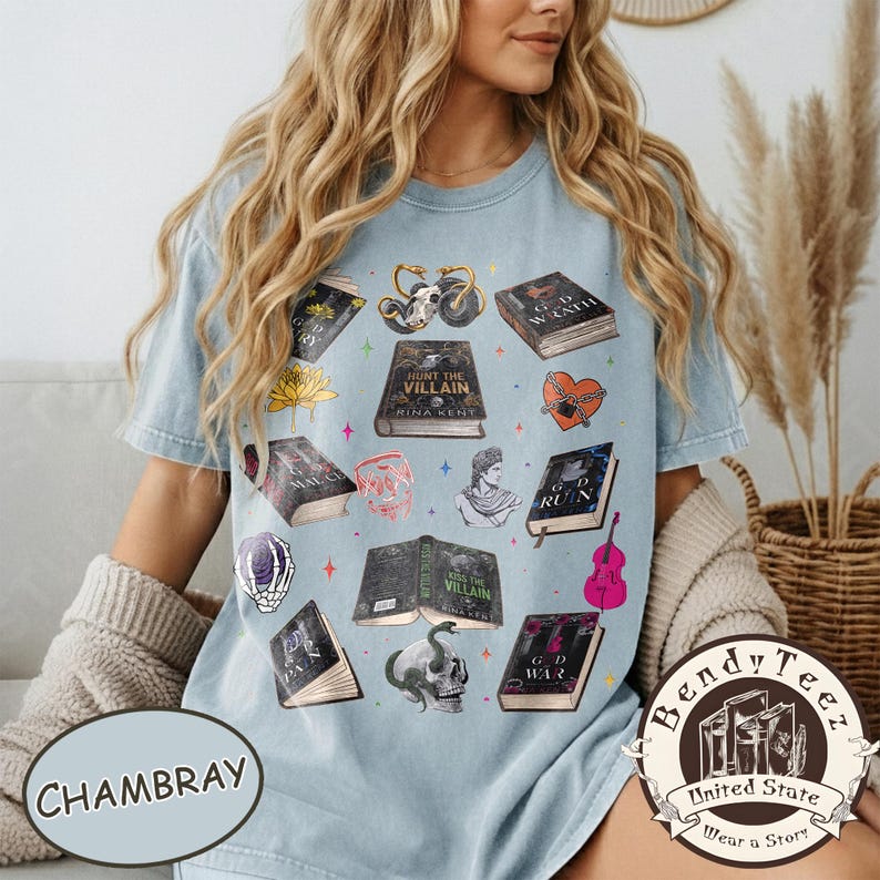 May include: Light blue chambray t-shirt featuring various book illustrations with titles like "Hunt the Villain" and "G.D. Wrath." The shirt also includes a skull, heart, and other graphics. The shirt has the text "Bendy Tee" and "United State Wear a Story".