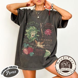 Peut inclure: T-shirt gris foncé avec un motif graphique représentant Méduse, une fleur de lotus et d'autres illustrations. Le t-shirt comprend du texte tel que "Aunty M's", "Lotus", "Blackjack" et "Delphi". Le t-shirt a un aspect vintage et vieilli.