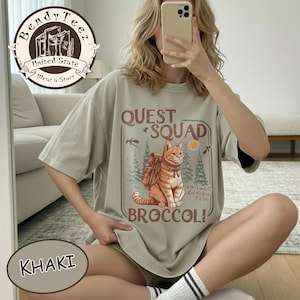 Könnte beinhalten: Khaki-farbenes T-Shirt mit einer Grafik, die eine Katze mit Rucksack, die Worte "QUEST SQUAD" und "BROCCOLI" zeigt. Das Design umfasst Bäume, Vögel und eine Sonne. Das Shirt hat kurze Ärmel.