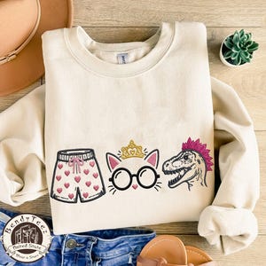 May include: A cream-colored crewneck sweatshirt featuring an embroidered design of a pair of shorts with pink hearts, a cat face wearing a gold crown and black glasses, and a dinosaur head with a pink mohawk, all centered on the front chest.
