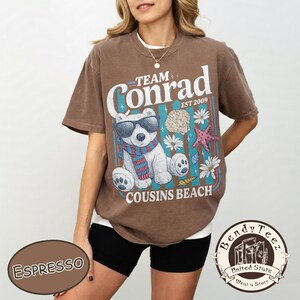 May include: Espresso-coloured t-shirt with a graphic featuring a polar bear wearing sunglasses and a scarf. The text reads "Team Conrad EST 2009 Cousins Beach" with seashell, starfish, and flower illustrations. The shirt is a relaxed fit.