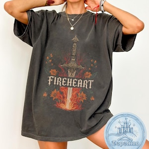 Puede incluir: Una camiseta gris carbón de manga corta con un gráfico central de una espada ornamentada rodeada de llamas naranjas y rojas y elementos florales. La palabra FIREHEART está impresa en una fuente serif negrita sobre el diseño de la espada.