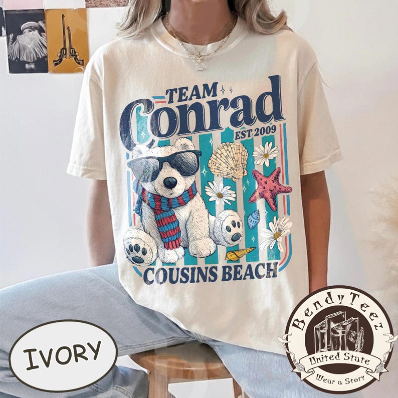 May include: Ivory t-shirt with a graphic of a bear in sunglasses and a scarf, with the text "Team Conrad Cousins Beach Est. 2009." The design includes seashells, starfish, and flowers. The shirt has a relaxed fit.