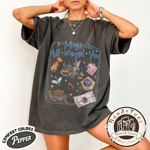 Puede incluir: Camiseta gris oscuro con el texto "Magic is All around You" y varios gráficos, incluyendo un castillo, una mariposa y botellas de pociones. La camiseta tiene un aspecto vintage y desgastado y presenta la marca Comfort Colors.
