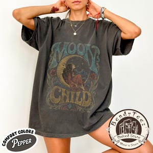 Könnte beinhalten: Ein anthrazitfarbenes Kurzarm-T-Shirt mit einem Grafikdruck, der eine Person auf einer Mondsichel zeigt, sowie dem Text "MOON CHILD" in einer stilisierten Schriftart. Das Design enthält florale Elemente, Wolken und Sterne in gedeckten Farben.