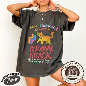 May include: Dark grey t-shirt with the text "BEING TRANSGENDER IS NOT A PERSONAL ATTACK" and a cat holding a rainbow flag. The shirt also has the text "(But if you keep being a bigoted asshole, things might get hairy)".