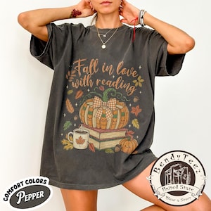 May include: A charcoal gray short-sleeve t-shirt featuring the text Fall in love with reading. The graphic includes a stack of books, a pumpkin with a plaid bow, a mug with a maple leaf, and autumn leaves. The shirt has a relaxed, loose fit.
