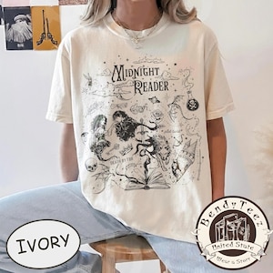 May include: An ivory short-sleeve t-shirt featuring a black graphic print with the text Midnight Reader, Death by TBR, and plot twist. The design includes illustrations of a book, a sword, a skull, a rose, a castle, and various fantasy elements.
