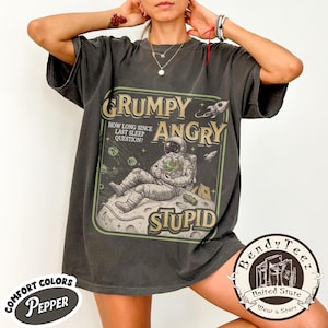 May include: Dark gray t-shirt with a distressed graphic featuring an astronaut on a moon, with the words "GRUMPY, ANGRY, STUPID." The shirt also has the text "HOW LONG SINCE LAST SLEEP QUESTION?" and "COMFORT COLORS PEPPER."