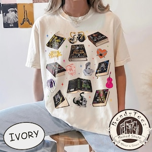 May include: Ivory-colored t-shirt featuring various book illustrations, including titles like "Hunt the Villain" and "God of Ruin." The design incorporates skulls, a violin, and other gothic-themed elements. The shirt also has the text "Bendy Teez" and "Wear a Story."