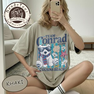 May include: Khaki t-shirt with a graphic of a polar bear wearing sunglasses and a scarf, with the text "Team Conrad Cousins Beach Est 2009". The shirt has short sleeves and a relaxed fit.