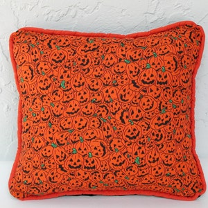 K&ouml;nnte beinhalten: Ein orangefarbenes Kissen mit Halloween-Motiv, das ein sich wiederholendes Muster aus schwarzen K&uuml;rbisgesichtern auf schwarzem Hintergrund zeigt. Das Kissen hat einen roten Rand.