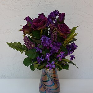 May include: A bouquet of purple roses and lilac flowers in a iridescent glass vase with swirling patterns.