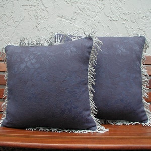 May include: Two blue throw pillows with a white fringe border and a floral pattern. The pillows are sitting on a wooden bench.