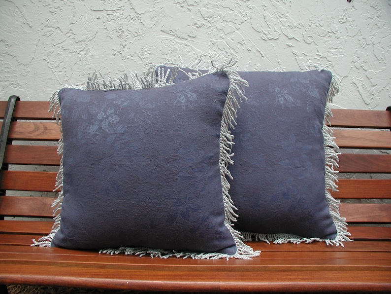 May include: Two blue throw pillows with a white fringe border and a floral pattern. The pillows are sitting on a wooden bench.