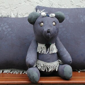 May include: A plush, gray teddy bear with a white beaded necklace and a white bow on its head. The bear is sitting on a wooden bench with two gray pillows with fringe trim.