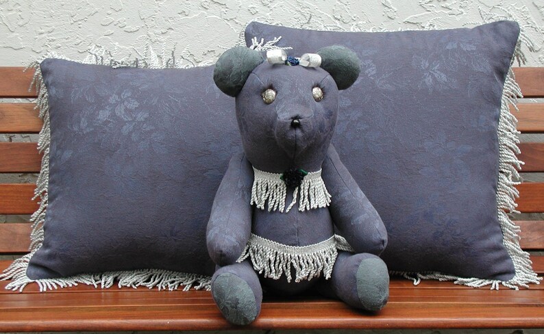 May include: A plush, gray teddy bear with a white beaded necklace and a white bow on its head. The bear is sitting on a wooden bench with two gray pillows with fringe trim.